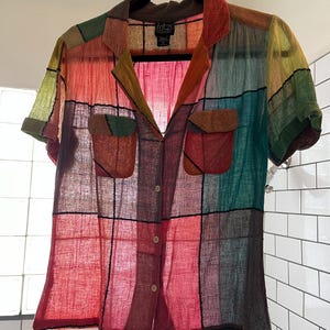 May include: A short-sleeved button-down shirt featuring a multi-colored patchwork grid pattern in shades of red, green, yellow, and blue. The shirt has a collar, two front chest pockets with flaps, and light-colored buttons. The label reads Fritzi.
