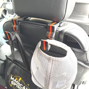 May include: Two black car seat headrest hooks with orange accents. A black tote bag with the text "Magical Spirits" hangs from one hook. A gray mesh baseball cap hangs from the other hook.