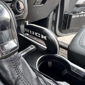 May include: Interior view of a vehicle's console, featuring a black and silver gear shift with a horse emblem and an American flag. A black and white "BUCK" handle is visible, along with a cup holder and a seat.