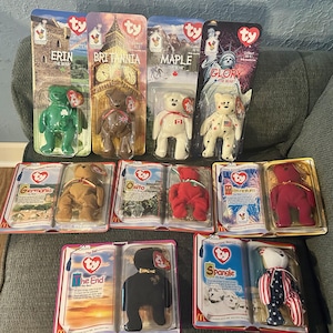 May include: A selection of Ty Beanie Babies, such as Erin, Britannia, Maple, and Glory, displayed in their original packaging. The plush toys are in various colours, some with patriotic designs. The packaging features the Ty heart logo.