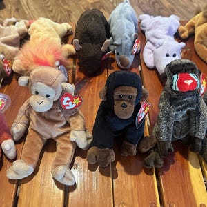 May include: A collection of plush animal toys, including monkeys, a gorilla, a sloth, and other creatures. The toys are in various colors and sizes, with tags attached. They are arranged on a wooden surface.
