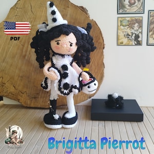May include: A crocheted doll dressed as a Pierrot, wearing a white and black costume with a pointed hat. The doll has black hair and is holding a small basket. The doll is standing on a wooden surface. The text "Brigitta Pierrot" is written below the doll.