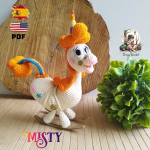 Puede incluir: Un caballito balancín de unicornio tejido a mano en crema, naranja y azul. El unicornio tiene un cuerno de arcoíris, una nariz rosa y detalles de estrellas y corazones. El caballito está sobre balancines de madera. La palabra "MISTY" está abajo.