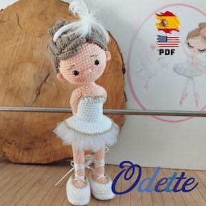 Puede incluir: Una muñeca bailarina de crochet con un tutú blanco, cabello castaño y zapatos blancos. La muñeca sostiene una pluma blanca en su cabello. La muñeca está de pie sobre una superficie de madera. El texto "Odette" está escrito en cursiva azul en la parte inferior derecha de la imagen.