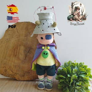 Könnte beinhalten: Handgefertigte Häkelpuppe mit grauem Hut, Draht- und roten Perlen-Heiligenschein, gelbem Hemd mit Alien-Grafik, lila Umhang und dunkelgrünen Shorts. Die Puppe hat eine blaue Augenklappe und steht vor einer Holzplatte.
