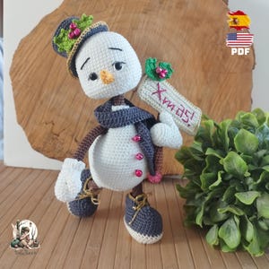 Puede incluir: Muñeco de nieve de ganchillo con bufanda, sombrero y zapatos azules. El muñeco de nieve sostiene un letrero que dice "Xmas!". Tiene nariz de zanahoria y botones rosas. Decoración navideña festiva.