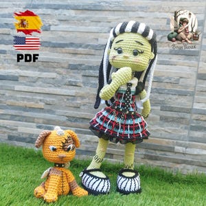 May include: Crocheted doll of a green-skinned character with black and white hair, wearing a plaid dress and black shoes. A small crocheted dog is also present. The dolls are set on green grass in front of a stone wall.