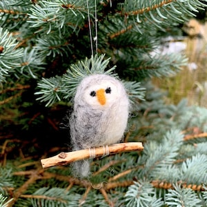 May include: A handmade owl ornament made of gray and white felt, perched on a small wooden branch. The owl has a yellow beak and black eyes. The ornament is hanging from a thin string against a backdrop of green pine needles.