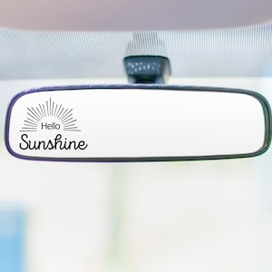 May include: A black rearview mirror with a white sticker that says "Hello Sunshine" with a sun graphic.