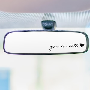 Pode incluir: Espelho retrovisor de carro com a frase "give 'em hell" em escrita cursiva, seguida por um símbolo de coração preto. O espelho é retangular com cantos arredondados e uma moldura preta, contra um fundo desfocado.