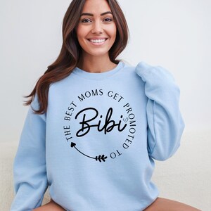 May include: Light blue sweatshirt with the text "The Best Moms Get Promoted to Bibi" in a black circular design. The word "Bibi" is in a cursive font. The sweatshirt is a solid light blue color.