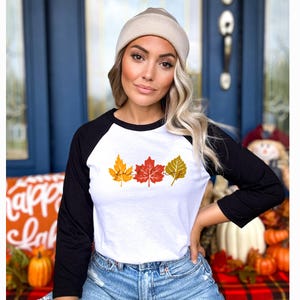 Op de afbeelding: Een wit raglan shirt met zwarte mouwen met drie kleurrijke herfstbladeren. De bladeren zijn geel, rood en goudkleurig, met het woord "fall" in een scriptlettertype. Het shirt wordt door een persoon gedragen.