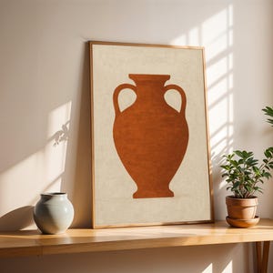 May include: A framed art print featuring a terracotta-colored vase on a cream background. The artwork is displayed on a wooden table, with a small gray vase and potted plant nearby. The frame has a gold finish.