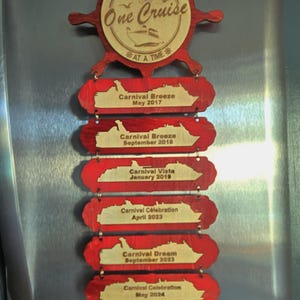 May include: A wooden ship wheel ornament with the text "ROLES FAMILY MAKING MEMORIES One Cruise AT A TIME" and a series of wooden plaques with the text "Carnival Breeze May 2017", "Carnival Breeze September 2018", "Carnival Vista January 2019", "Carnival Celebration April 2023", "Carnival Dream September 2023", "Carnival Celebration May 2024", and "Carnival Freedom September 2024".