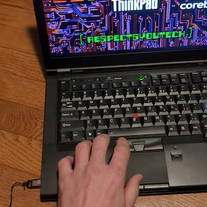 May include: A black laptop computer with a bright, colourful screen displaying the text "Lenovo ThinkPad + coreboot [RESPECTSYOUTECH]" on a circuit board background. The laptop is on a wooden surface with a hand resting on the touchpad.