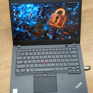 May include: A dark gray ThinkPad laptop with a screen displaying a digital lock icon. The keyboard has a red trackpoint and a touchpad. A USB drive is plugged into the side. The laptop is on a wooden surface.