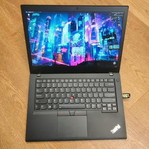May include: Black laptop with an open screen displaying a colorful cityscape with neon lights. The laptop has a full keyboard, a touchpad, and a red pointing stick. A USB drive is plugged into the side. The laptop is on a wooden surface.