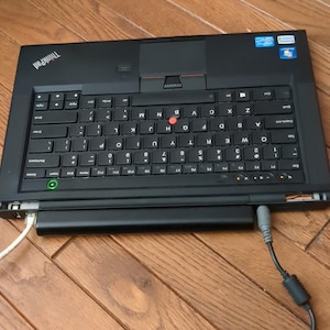 May include: A black ThinkPad laptop with a red dot on the keyboard. The laptop is open and the screen is facing away from the camera. The laptop is plugged into a power adapter.