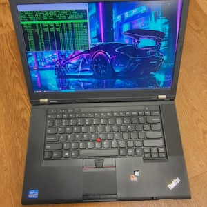 May include: Black ThinkPad laptop with a screen displaying a car graphic and lines of green text. The keyboard has a red trackpoint and a touchpad. The laptop is open on a wooden surface.