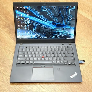 May include: A black ThinkPad laptop with an open screen displaying a blue and black abstract design. The keyboard has a red pointing stick and a touchpad. A USB drive is plugged into the side. The laptop is on a light brown wooden surface.