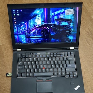 May include: A black Lenovo ThinkPad laptop with a QWERTY keyboard, a red pointing stick, and a trackpad. The screen displays a vibrant blue and purple digital wallpaper of a sports car in a city street, with a small sticker in the top right corner.
