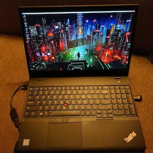 May include: A black Lenovo ThinkPad laptop with a full QWERTY keyboard, a red pointing stick in the center, and a numeric keypad. The screen displays a vibrant digital cityscape wallpaper with red and blue neon lights and a central figure.