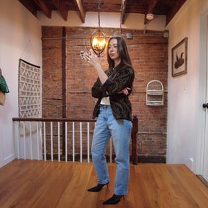 May include: A woman wearing a brown and black patterned jacket, blue jeans, and black heeled shoes stands in a hallway with exposed brick walls.