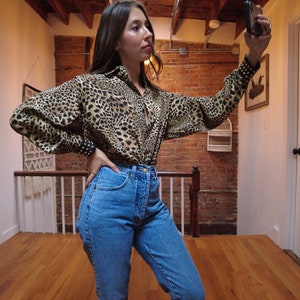 May include: A woman wearing a leopard print button-up shirt with black studded cuffs and blue denim jeans.