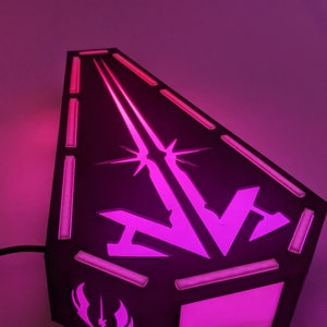 May include: A black light-up lamp shaped like a spaceship with a pink light emanating from the inside. The lamp features a stylized spaceship design with a starburst and a symbol of a lightsaber and wings on the bottom.