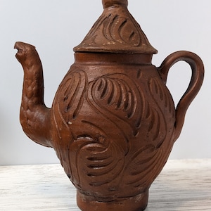 May include: A brown clay teapot with a lid and a spout shaped like a bird's head. The teapot is decorated with a carved floral pattern.