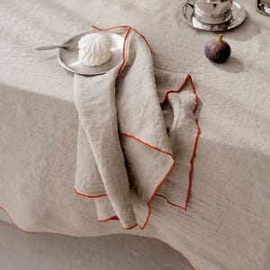 May include: A table setting with a neutral linen tablecloth, featuring silver cups and saucers, a white bowl of meringues, fresh figs, and a folded linen napkin with red trim. The scene evokes a sense of elegance and simplicity.