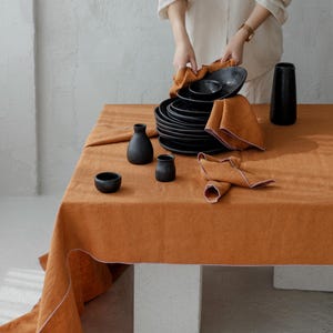 May include: A table setting featuring a burnt orange linen tablecloth with white trim. The table is set with black plates, bowls, napkins, and small black vases. The setting is on a white surface.