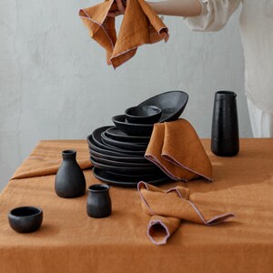 May include: An arrangement of black ceramic tableware, including plates, bowls, and vases, set on a rust-colored linen tablecloth. Two folded orange napkins with pink trim are visible. One napkin is being held up.