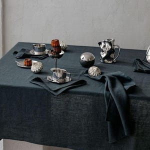 Può includere: Una tavola apparecchiata con una tovaglia di lino blu scuro. La tavola è apparecchiata con tazze, piattini, piattini e oggetti decorativi argentati. Sono presenti anche tovaglioli blu scuro e piccole torte.