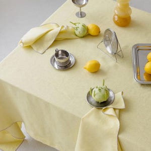 May include: A pale yellow tablecloth is set with lemons, napkins, and serving pieces. A silver tray holds lemons and a green fruit. A stack of glass spheres and a small silver cup and saucer are also on the table. The scene is brightly lit.
