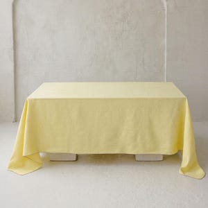 May include: A pale yellow tablecloth, rectangular in shape, covering a table. The fabric has a soft, textured look and drapes down on all sides. The background is a light-coloured wall with an arched architectural feature.