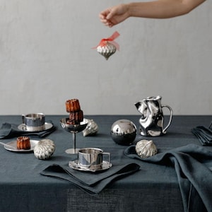 Puede incluir: Una mesa con un mantel azul oscuro presenta tazas, platillos y adornos decorativos plateados. Pequeños pasteles están en platos y en un plato plateado. Una mano sostiene un adorno plateado con una cinta roja.
