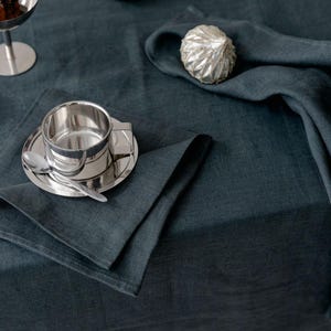 Puede incluir: Un mantel de lino verde azulado oscuro con un juego de taza y platillo de plata, una cuchara y una servilleta doblada. También hay esferas decorativas plateadas y un pequeño objeto plateado sobre la mesa. Un pequeño postre está en una copa de plata.