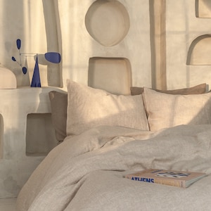 May include: A beige linen duvet cover with two beige linen pillows and a book titled "ATLiens" on top. The bed is in a room with a white wall with several rectangular and arched niches.