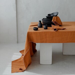 May include: A table setting featuring a burnt orange linen tablecloth. Black ceramic plates, bowls, and small vases are arranged on the table. Two folded napkins and a rolled napkin are also present. The table is supported by white rectangular blocks.