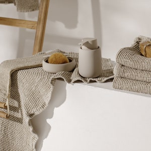 Könnte beinhalten: Ein Set an Badezimmer-Essentials in einer neutralen Farbpalette. Das Set beinhaltet einen beigefarbenen Seifenspender, eine beige Schale mit einem braunen Schwamm und einen Stapel beigefarbener Waffel-Handtücher.