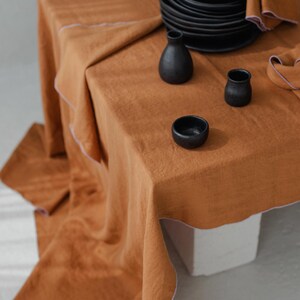 May include: A table setting featuring a burnt orange linen tablecloth with a white trim. Black ceramic plates, bowls, and small vases are arranged on the table. A folded napkin with a pink trim is also present.
