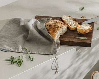 Puede incluir: Una bolsa de pan de lino gris con cierre de cordón, sentada sobre una encimera blanca junto a una tabla de cortar de madera con pan rebanado y hojas de salvia.
