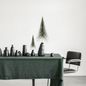 Puede incluir: Un mantel de lino verde oscuro cubre una mesa con patas de metal negro. Una fila de pequeños jarrones negros y un jarrón negro más grande con un pequeño árbol de Navidad artificial verde están dispuestos sobre la mesa. Una silla negra con patas cromadas se encuentra a la derecha de la mesa.