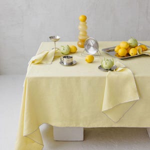 May include: A yellow tablecloth with a matching napkin and silver serving pieces. Lemons and green artichokes are arranged on the table. A decorative glass vase with stacked lemons is also present. The table is set against a neutral background.