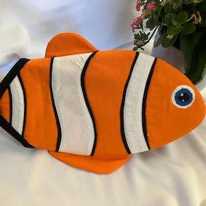 May include: An orange and white fish-shaped pillow with black trim and a blue eye. The pillow has a tail and fins, and is designed to resemble a clownfish. The pillow is on a white surface.