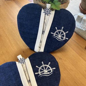 May include: Two navy blue heart-shaped items with white accents and nautical embroidery. Each has a white stripe down the center and a small ship wheel charm. The embroidery features a ship's wheel design.