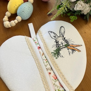 May include: White, heart-shaped pot holder with a floral pattern and a bunny embroidered on the front, holding carrots. The pot holder has a lace trim and a loop for hanging. Easter-themed.