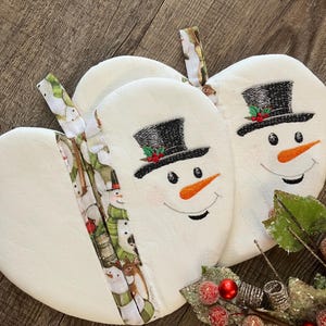 May include: White snowman-shaped quilted pot holders with black top hats, orange carrot noses, and embroidered smiles. The edges are trimmed with a patterned fabric featuring snowmen and winter scenes. A festive holiday kitchen item.