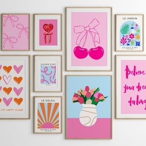 May include: A gallery wall with nine framed prints featuring various designs. The prints include hearts, cherries, flowers, a sun, and text. The prints are all in pink, orange, red, blue, and white.